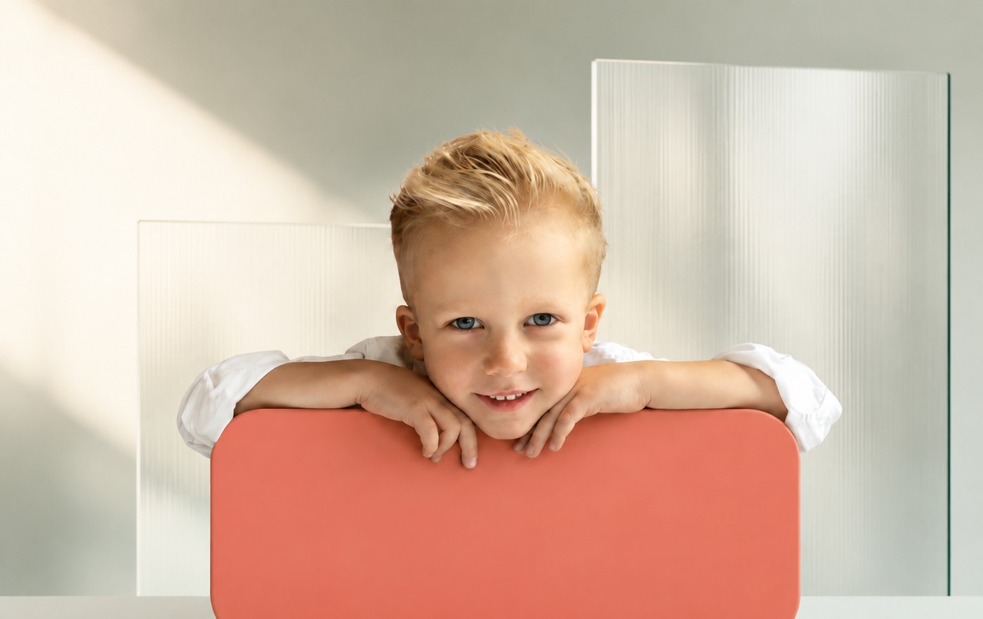A young boy leaning on a coral panel, smiling at the camera, with frosted glass panels behind him.